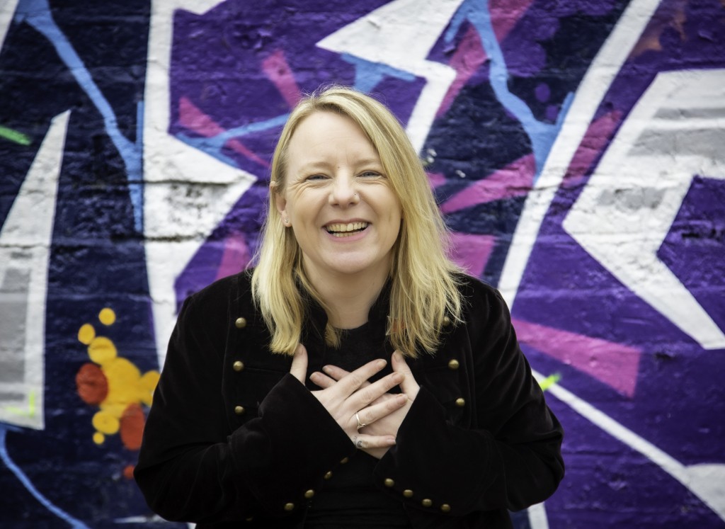 A picture of Awen Clement, a white woman who is standing in front of a colourful graffiti wall. She is smiling and has her hands pressed to her chest. 